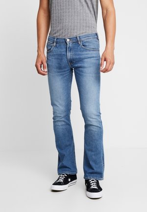 Mężczyzna w jasnoniebieskich dopasowanych jeansach bootcut, szarym bezrękawniku z prążkowanej dzianiny oraz czarnych niskich trampkach z białymi sznurówkami i podeszwami.