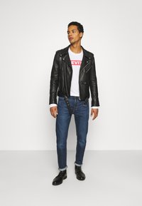 Giacca da moto in pelle nera sopra una maglietta bianca con logo rosso, abbinata a jeans blu e stivali neri con lacci, con maniche arrotolate.