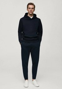 Námornícky modrá mikina s kapucňou a zodpovedajúce úzke tepláky z mäkkej tkaniny. Model má na sebe biele tenisky. Jednoduchý dizajn, žiadne viditeľné vzory alebo logá.