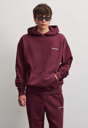 Ung mand iført mørkerød hættetrøje og matchende joggingbukser med PEGADOR-logo, poserer med den ene hånd i lommen og solbriller.