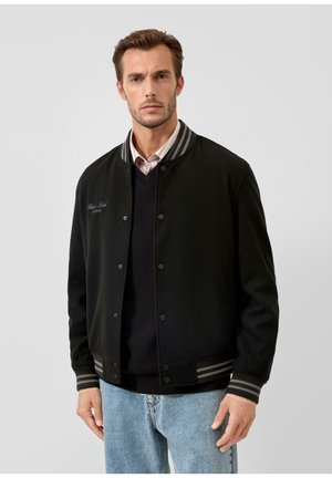 Man draagt een zwarte varsityjas met gestreepte manchetten en kraag, gecombineerd met een zwarte trui en lichtblauwe spijkerbroek, staande tegen een effen achtergrond.