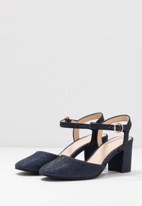 Blå glittrande klackar med spetsig tå, ankelsrem med rund spänne och klack med blockform; slät textur med glittrigt avslut.