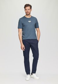 Niebieska koszulka z krótkim rękawem z białą łatką logo, w połączeniu z granatowymi spodniami i białymi sneakerami. Model stoi z rękami w kieszeniach.