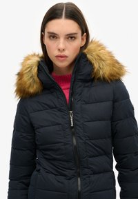 Marineblauwe gewatteerde winterjas met een met bont gevoerde capuchon, voorzien van een ritssluiting aan de voorkant, een gekwilt textuur en een logo-detail op de mouw.