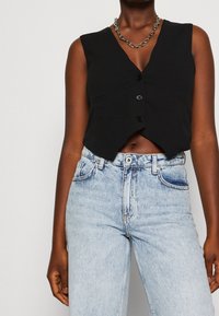 Person som bär en ärmlös svart knäppt väst, ljusblå högmidjade jeans och ett kraftigt silverfärgat kedjehalsband mot en neutral bakgrund.