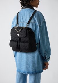 Black quilted backpack with chain detail, two front pockets, and a top flap. Adjustable straps and contrasting silver hardware.