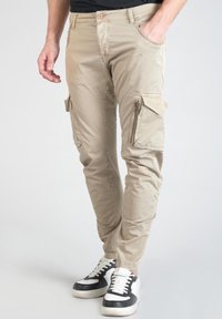 Beige cargobroek met een slanke pasvorm, voorzien van ritszakken en een stevige stof. Gepaard met zwart-witte sneakers.