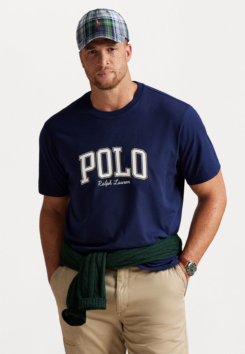Sötétkék pamut póló "POLO Ralph Lauren" logóval, derékra kötött zöld kötött pulóverrel és kockás sapkával párosítva. Khaki nadrág.