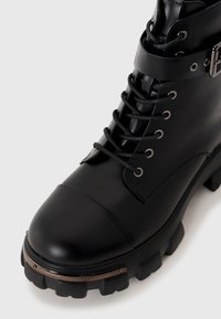 Black lace-up ankle boot made of smooth, glossy material with a rounded toe and rugged sole; features a metal accent and buckle detail.