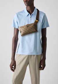 Braune gemusterte Umhängetasche mit goldenem Reißverschluss an einem Modell, das ein hellblaues Poloshirt und beige Hosen trägt und ein schlankes Design zeigt.