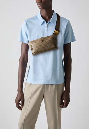 Borsa a tracolla marrone a fantasia con zip dorata su un modello che indossa una polo azzurra chiara e pantaloni beige, che mette in mostra un design elegante.