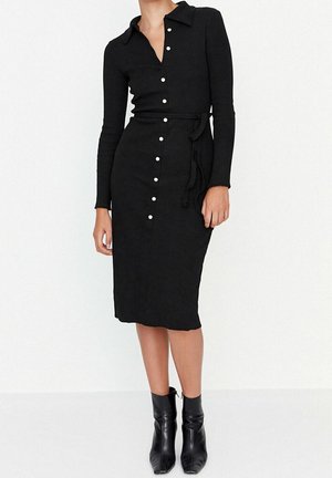 Femme portant une robe midi noire ajustée à manches longues avec des boutons blancs sur le devant et des bottines noires sur un fond uni.