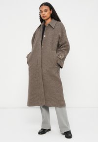 Brun og beige ternet oversized frakke med klassisk krave, knaplukning og sidelommer, lagd over grå bukser.