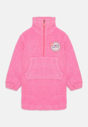 Roze fleecejurk met een hoge kraag, halve rits, voorzak en een decoratieve patch van een glimlachend gezicht met hartjes en bloemen.