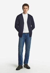 Marinblå cardigan med V-ringning, knappframsida och ribbade ärmslut, parat med en vit T-shirt och blå denimjeans. Bruna skor fulländar looken.