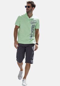 Helles grünes Polohemd mit bedrucktem Text auf der Vorderseite, kombiniert mit dunkelgrauen Cargo-Shorts und weißen Sneakers, die vor einem schlichten Hintergrund gehen.