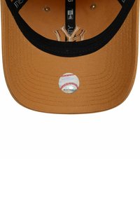 Gorra de los New York Yankees con un cuerpo de algodón beige, visera negra y una pegatina circular de béisbol en la parte inferior. Presenta costuras decorativas.