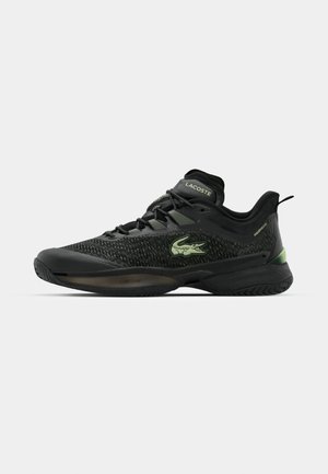 Zapato deportivo negro con parte superior de malla texturizada, logo verde y suela de goma. Presenta cordones y un talón acolchado para mayor soporte.