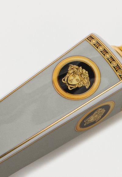 Harmaa suorakaiteen muotoinen säiliö, jossa on kultainen ja musta Medusan pää -embleemi sekä koristeellinen kultainen reunus valkoisella taustalla.