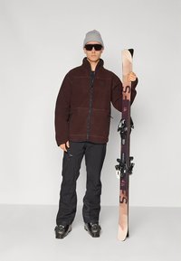 Pruun fliisjakk tõmblukuga, mustad püksid, suusaapad ja päikeseprillid. Käes suusad musta kinnitusdetaili ja helepruunide otstega. Hall müts.