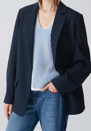 Kobieta w granatowej marynarce na białym i niebieskim pasiastym topie bez rękawów oraz niebieskich dżinsach, z jedną ręką w kieszeni.