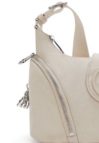 Borsa a mano beige chiaro con una superficie liscia, dotata di una cerniera e accessori in argento, tracolla regolabile e un charm decorativo.