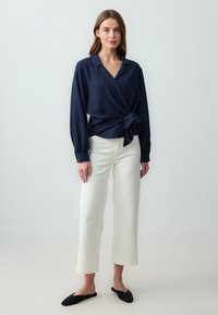 Blusa avvolgente blu navy a maniche lunghe con vita legata, abbinata a pantaloni bianchi a gamba larga e scarpe nere slip-on. Tessuto dalla texture liscia.