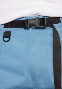 Svart plastspänne som fäster ett svart justerbart bälte över ljusblå byxor, bärs med en vit skjorta.