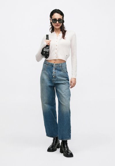 Fehér, gombos, rövid kardigán, világoskék, magasderekú, bő szárú farmernadrág, fekete masszív bokacsizma, fekete táskát tartva.