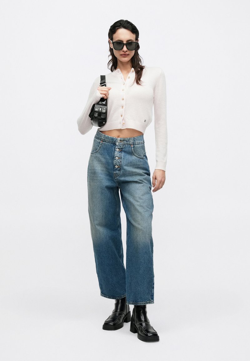 Weißes kurz geschnittenes Strickjacke mit Knopfleiste, hoch taillierte hellblaue Jeans mit weitem Bein und schwarze grob gearbeitete Stiefeletten, hält eine schwarze Tasche.