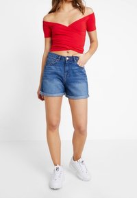 Kvinde iført rød off-shoulder cropped top, rullede denim shorts og hvide sneakers, stående mod en hvid baggrund.