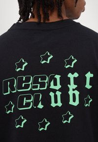 Black cotton t-shirt featuring a large green graphic text "RESORT CLUB" and star designs, showcasing a bold, playful font and color contrast.