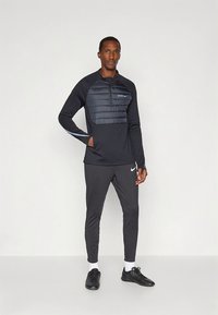 Top atletico nero con sezione imbottita sul petto, maniche lunghe e collo con cerniera. Abbinato a pantaloni neri aderenti e scarpe da ginnastica nere.