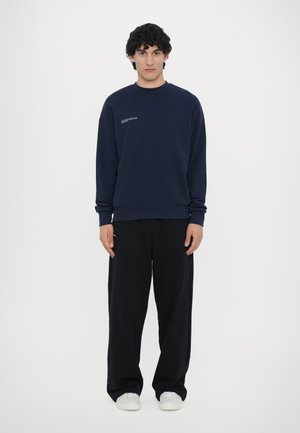 Mørkeblå sweatshirt med runde halsudskæring, ribbede manchetter og et lille grafik på brystet, parret med løse sorte bukser og hvide sneakers.
