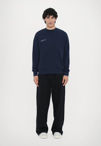 Navy blue sweatshirt with crew neck, ribbed cuffs, and a small graphic on the chest, paired with loose black trousers and white sneakers.