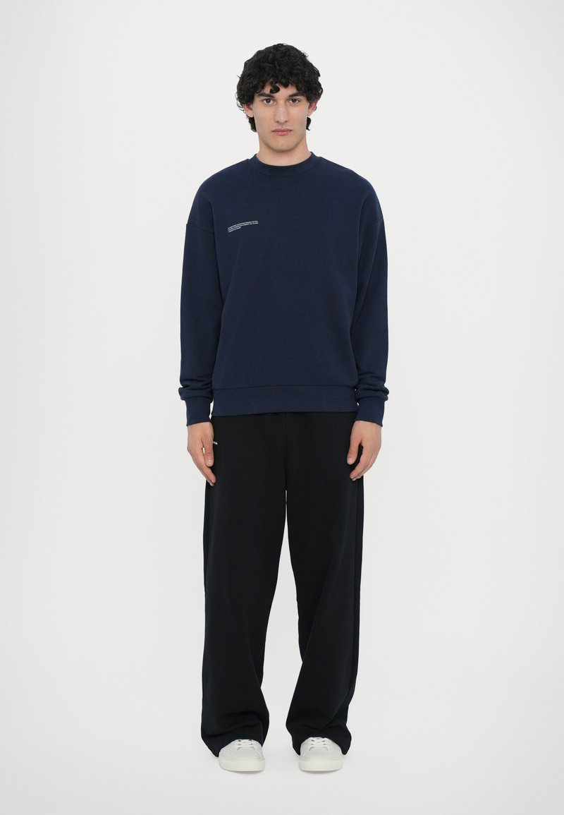 Mørkeblå sweatshirt med runde halsudskæring, ribbede manchetter og et lille grafik på brystet, parret med løse sorte bukser og hvide sneakers.