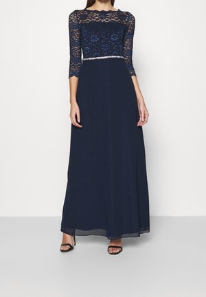 Kobieta ubrana w granatową suknię sięgającą podłogi z koronkowym gorsetem, rękawami do łokcia i cienkim srebrnym paskiem, do tego czarne sandały na wysokim obcasie.