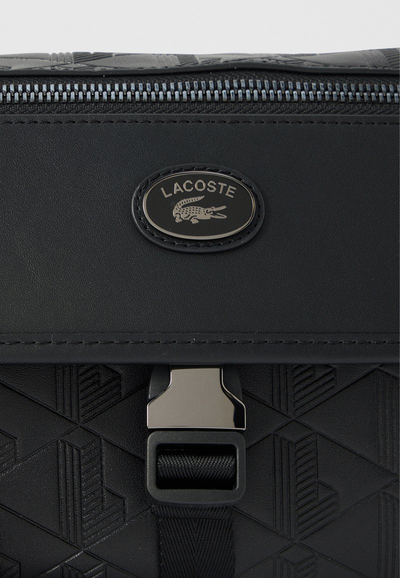Sort struktureret taske med geometriske mønstre, med en sølv Lacoste-logo, lynlås og en metallisk låsedetalje.
