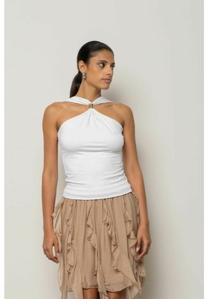 Donna con i capelli scuri legati indossa un top bianco a halter con fibbia dorata e una gonna beige a più strati con volant, guarda di lato.