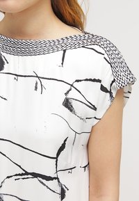 Blusa bianca con motivi astratti a pennellate nere, colletto tessuto a trama e maniche corte.