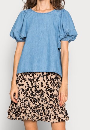 Mujer con una blusa de mezclilla azul claro con mangas abullonadas, combinada con una falda beige con volantes que presenta un estampado de leopardo en negro y marrón.