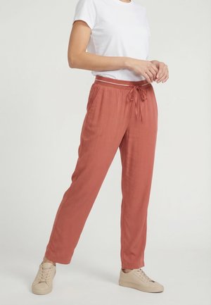 Donna che indossa pantaloni ruggine con coulisse, t-shirt bianca e sneakers beige, in piedi davanti a uno sfondo chiaro uniforme.