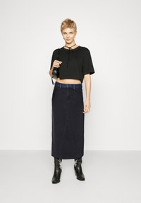 Zwarte cropped top met een gestrikt decolleté, gecombineerd met een zwarte denim maxi rok met een blauwe tailleband en voorkant stikseldetails. Zwarte laarzen.