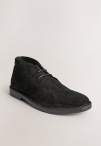 Bottine en cuir suédé noir avec lacets à l'avant et semelle basse en caoutchouc sur fond clair.