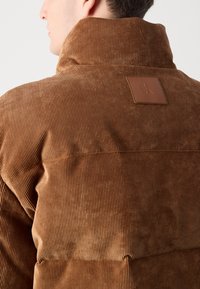 Braune Cordjacke mit hohem Kragen und einem Leder-Logo-Patch auf dem Rücken, das eine vertikale Rippstruktur zeigt.