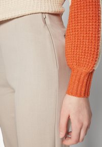 Pantalons beige ajustés avec une texture lisse, associés à un pull à manches en tricot côtelé orange présentant un motif contrasté.