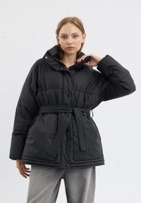 Doudoune noire avec un col haut, texture quiltée, fermeture à boutons-pression et à zip, dotée d'une taille ceinturée pour un ajustement réglable.