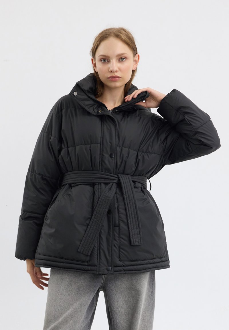 Doudoune noire avec un col haut, texture quiltée, fermeture à boutons-pression et à zip, dotée d'une taille ceinturée pour un ajustement réglable.