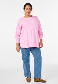 Różowa bluzka z długim rękawem i okrągłym dekoltem, zestawiona z niebieskimi jeansami zdobionymi perełkami i luźnym krojem. Brązowe baleriny.