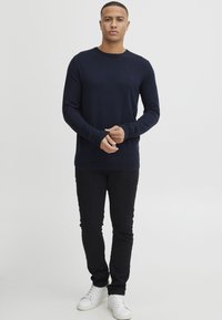 Hombre de pie vistiendo un suéter de manga larga azul marino, pantalones negros y zapatillas blancas frente a un fondo claro y liso.